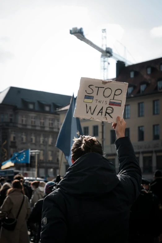 a person holding a sign that says stop war, a photo, trending on unsplash, viennese actionism, square, blue and yellow ribbons, nuremberg, 🚿🗝📝