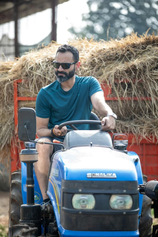 a man riding on the back of a blue tractor, navid negahban, stubble beard, wearing a dark blue polo shirt, bali