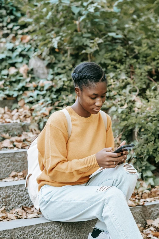 a woman sitting on steps looking at her cell phone, trending on pexels, black teenage boy, brown sweater, lush surroundings, yellow clothes