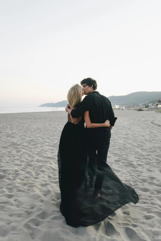 a couple of people standing on top of a sandy beach, wearing a black dress