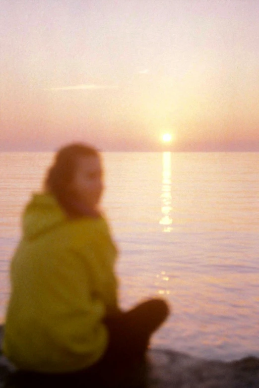 a person sitting on a rock in front of a body of water, video art, early morning sunrise, expired color film, yellow, calm face