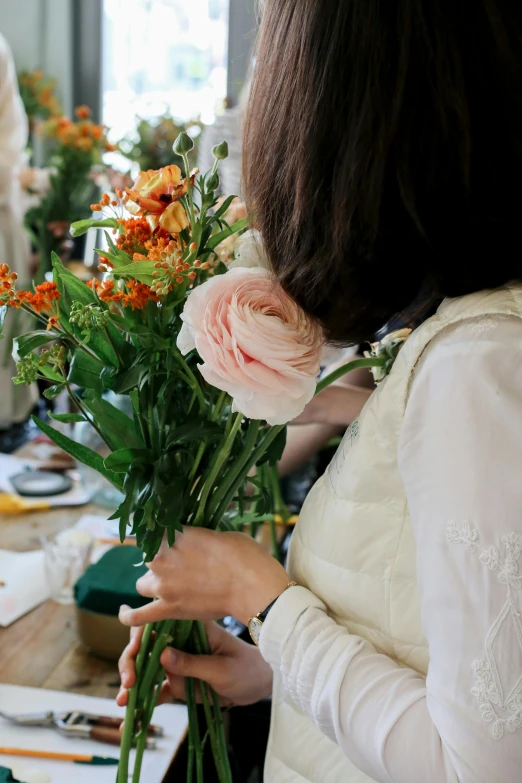 a woman holding a bunch of flowers at a table, inspired by Ruth Jên, carefully crafted, creating a soft, back - shot