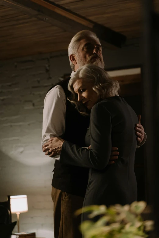 a man and a woman standing next to each other, embrace, silver hair and beard, standing in a dimly lit room, worksafe. cinematic