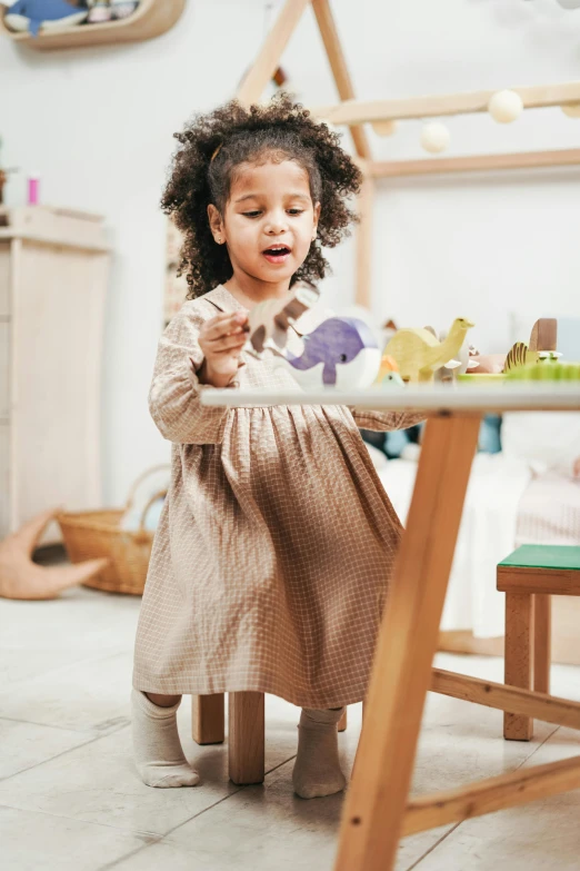 a little girl sitting at a table with a plate of food, inspired by Elsa Beskow, pexels contest winner, wooden art toys on base, walking to the right, dinosaur wooden statue, activity play centre