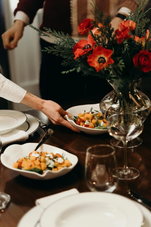 a group of people standing around a table with plates of food, with flowers, moody vibe, fine dining, salad