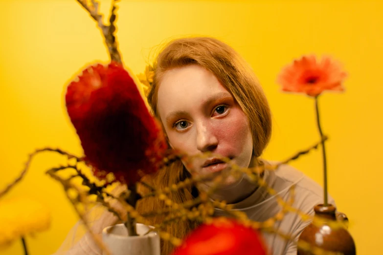 a woman sitting at a table with a vase of flowers, an album cover, inspired by Ignacy Witkiewicz, pexels contest winner, human face with bright red yes, sansa, showstudio, yellow and red