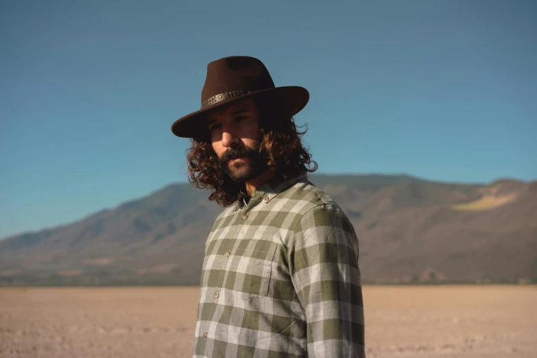 a man with long hair wearing a hat, inspired by Germán Londoño, unsplash, flannel, desert in the background, green and brown tones, california;