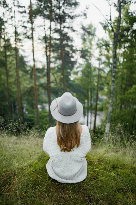 a woman sitting on top of a lush green field, by Anna Boch, pexels contest winner, white hat, bright nordic forest, back facing, grey