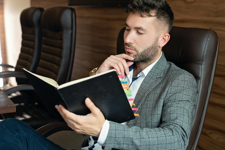 a man sitting in a chair reading a book, inspired by Nelson Alexander Ross, pexels contest winner, wearing business casual dress, multicoloured, holding pencil, attractive man