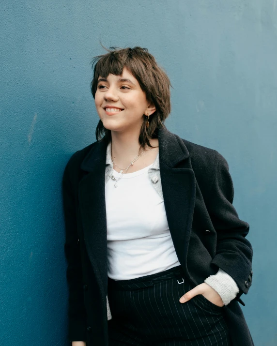 a woman standing in front of a blue wall, an album cover, by Grace Polit, wearing a black jacket, rebecca sugar, smiling and looking directly, profile image