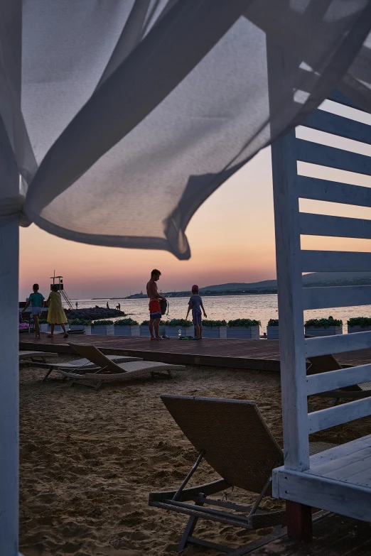 a white gazebo sitting on top of a sandy beach, during a sunset, people walking around, at the terrace, greek setting