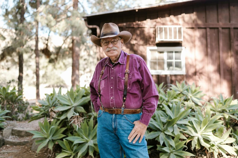 a man in a cowboy hat standing in front of a house, a portrait, by Leland Bell, unsplash, man with glasses, california;, he is at camp, 7 0 years old