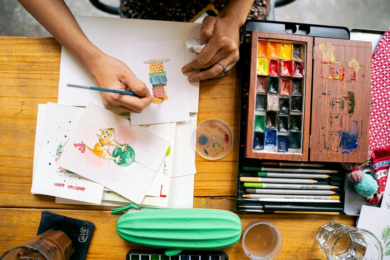 a wooden table topped with lots of art supplies, a watercolor painting, inspired by Jerry Pinkney, pexels contest winner, girl creates something great, 🎨🖌, stop motion animation, painting on a badge