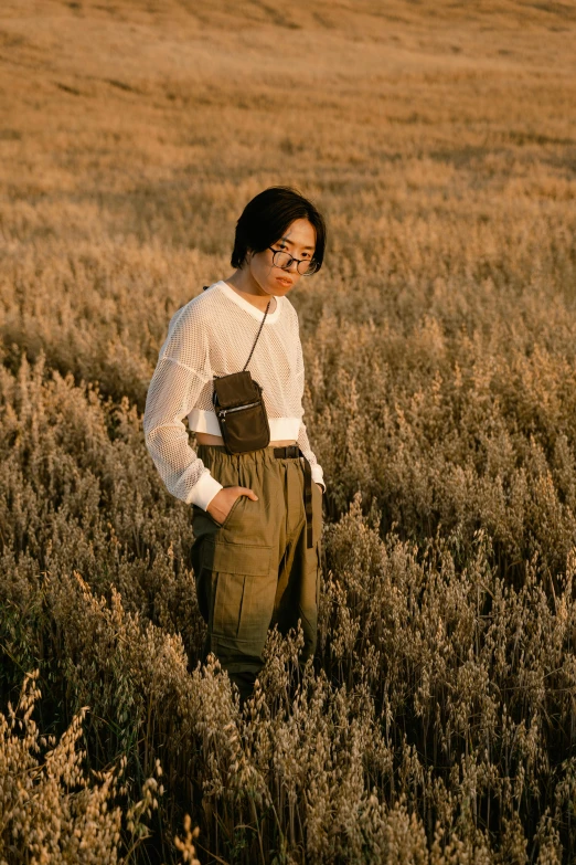 a man standing in a field of tall grass, inspired by jeonseok lee, trending on pexels, portrait androgynous girl, wearing cargo pants, small vials and pouches on belt, with square glasses