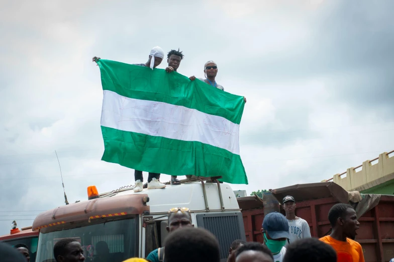 a group of people standing on top of a truck, a photo, trending on unsplash, happening, green flag, adebanji alade, flag in his right hand, square