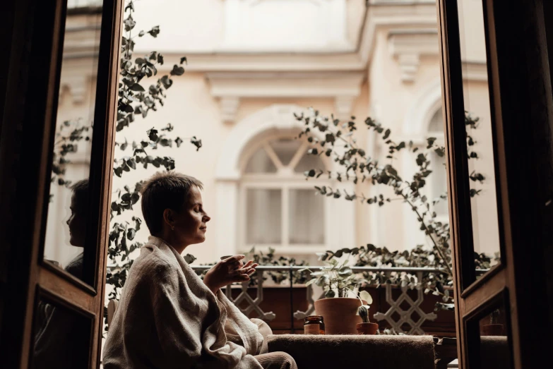 a woman sitting on a window sill next to a potted plant, by Emma Andijewska, pexels contest winner, visual art, cozy cafe background, profile image, sitting on a mocha-colored table, androgynous person