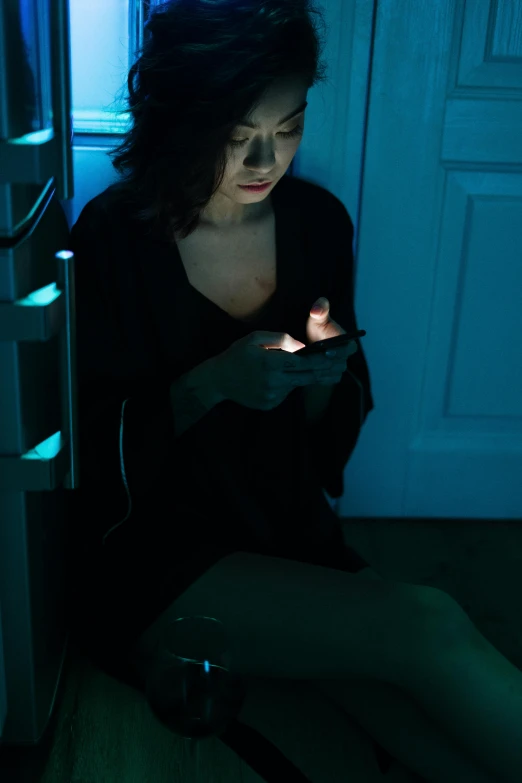 a woman sitting on the floor looking at her cell phone, inspired by Elsa Bleda, happening, horror movie lighting, dark teal lighting, woman in black robes, with blue light inside