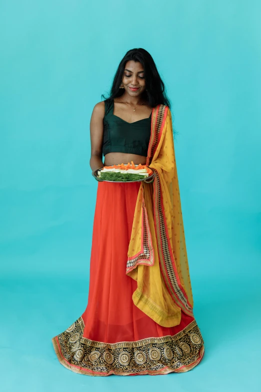 a woman holding a tray of food on a blue background, by Natasha Tan, hurufiyya, dressed in long fluent skirt, vibrant red and green colours, wearing yellow croptop, red silk flowing fabric