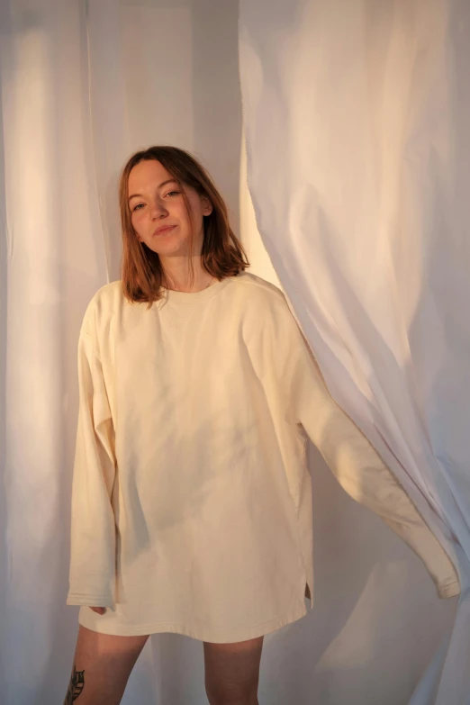 a woman standing in front of a white curtain, by Ellen Gallagher, wearing sweatshirt, simple cream dress, soft warm light, ilse gort