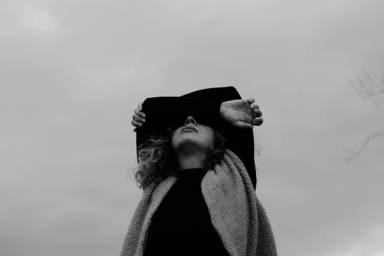 a black and white photo of a woman with her hands on her head, by Emma Andijewska, unsplash, surrealism, black stetson and coat, covered with blanket, low angle facing sky, dark visor covering face