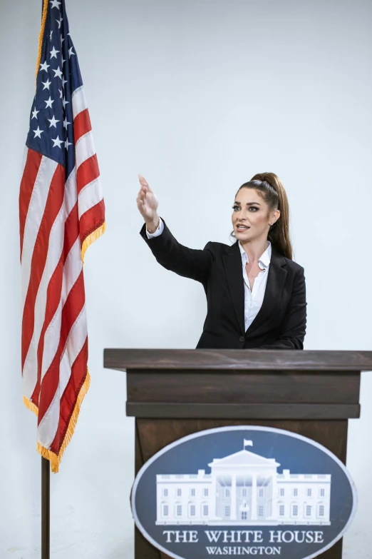 a woman giving a speech in front of an american flag, ashley greene, in the white house, pointing, promo image