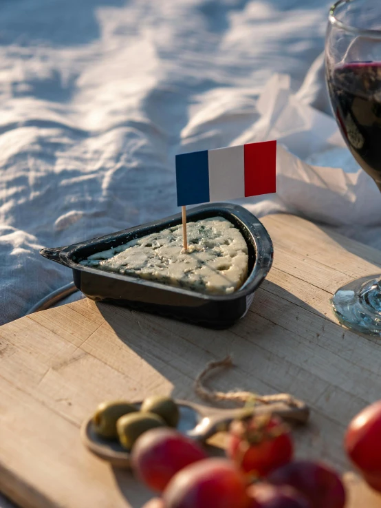 a wooden cutting board topped with cheese next to a glass of wine, french flag, slide show, alessio albi, seaside