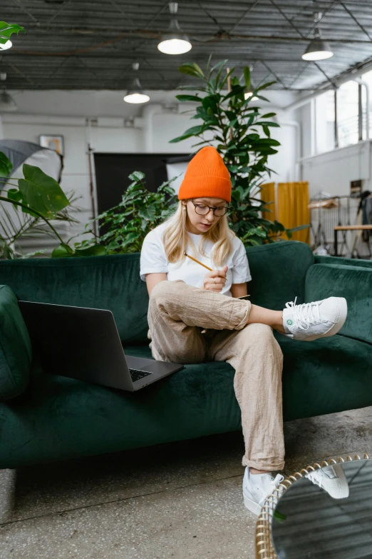 a woman sitting on a green couch with a laptop, trending on pexels, visual art, beanie, orange theme, wearing white sneakers, holding pencil