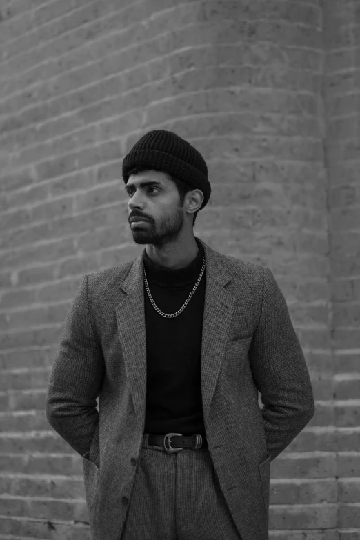 a man standing in front of a brick wall, a black and white photo, inspired by Theo Constanté, a portrait of rahul kohli, wearing a turtleneck and jacket, he is wearing a hat, drew hill