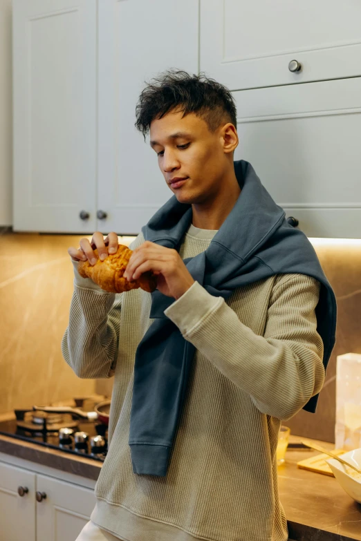 a man standing in a kitchen holding a sandwich, model wears a puffer jacket, scarf, wearing nanotech honeycomb robe, eating garlic bread