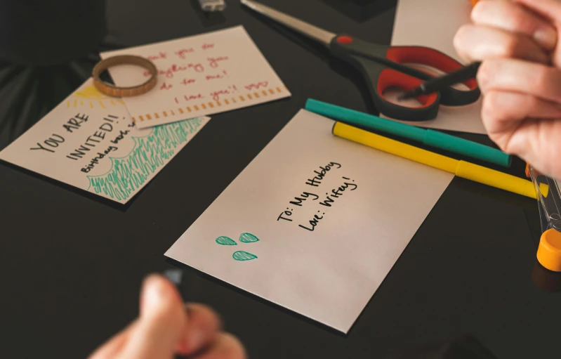 a person cutting a piece of paper with a pair of scissors, by Julia Pishtar, pexels contest winner, letterism, greeting card, back of hand on the table, sharpie, black and teal paper