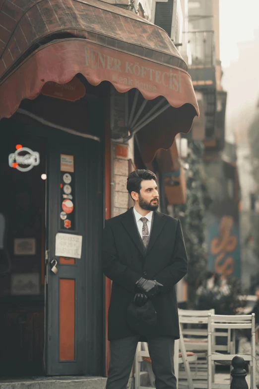 a man in a suit and tie standing on a sidewalk, an album cover, inspired by Ramon Pichot, pexels contest winner, renaissance, fallout style istanbul, standing in a restaurant, black overcoat, bearded