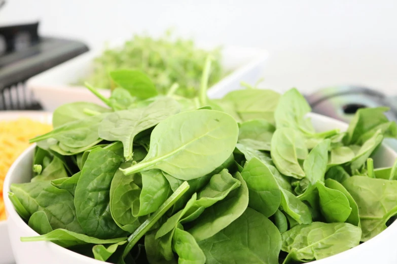 a bowl of spinach sitting on top of a counter, a picture, by Nicolette Macnamara, pexels, with a white background, closeup at the food, green: 0.5, looking from shoulder