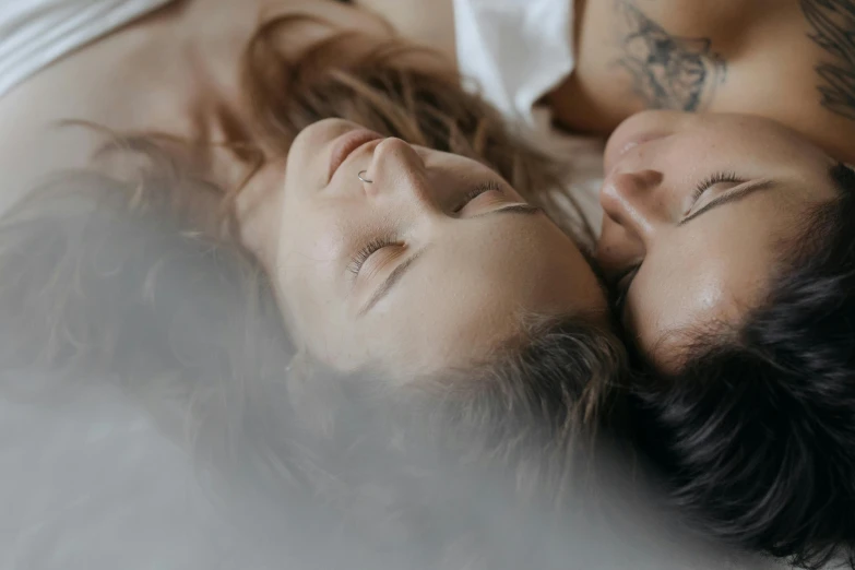 a couple laying on top of a bed next to each other, a tattoo, trending on pexels, lesbian embrace, dewy skin, gif, a woman's face