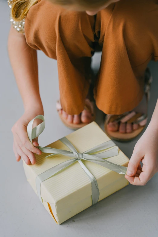 a little girl sitting on the floor holding a present, by Eden Box, pexels contest winner, renaissance, yellow and olive color scheme, minimalist, ribbon, sustainable materials