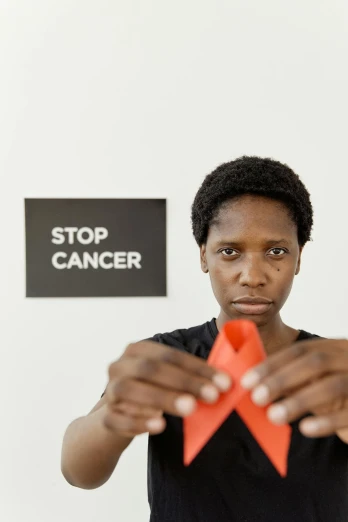 a woman holding a red ribbon in front of a stop cancer sign, a photo, by Lily Delissa Joseph, pexels contest winner, renaissance, ( ( dark skin ) ), caspar david, helio oiticica, stop motion