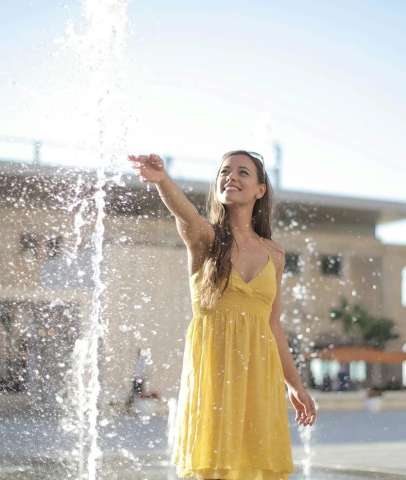 a woman in a yellow dress standing next to a water fountain, pexels, happening, being delighted and cheerful, realflow, sunshine, square