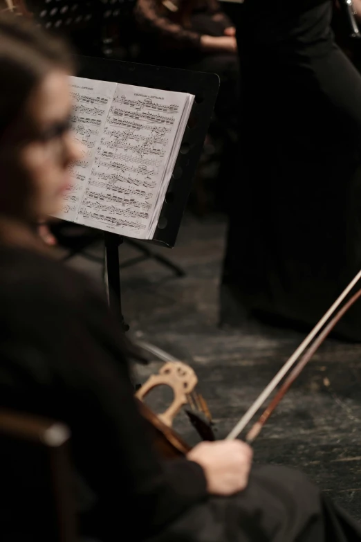 a woman in a black dress playing a violin, photo still of behind view, sheet music, square, audience