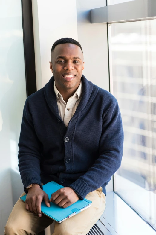 a man sitting on a window sill holding a book, jamal campbell, in a navy blue sweater, in the office, professional profile picture