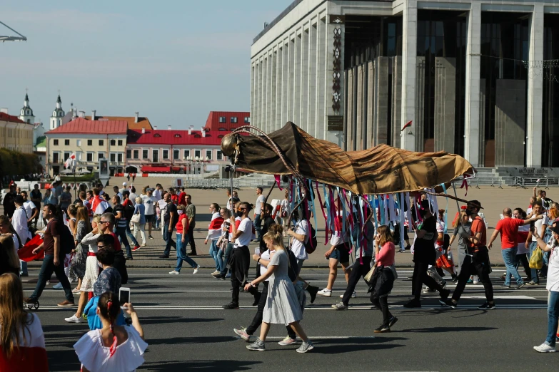 a crowd of people walking across a street, an album cover, by Grytė Pintukaitė, pexels contest winner, socialist realism, magic parade float, big long cloth on the wind, neo norilsk, a creature 5 meters tall