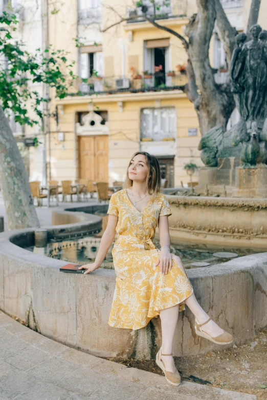 a woman sitting on a wall next to a fountain, inspired by Valéria Dénes, trending on pexels, wearing a yellow dress, town square, patterned clothing, lourmarin