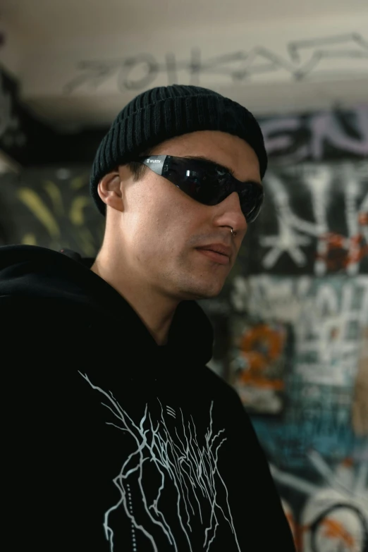 a man standing in front of a wall covered in graffiti, wearing glack glasses, tony hawk, with black beanie on head, **cinematic