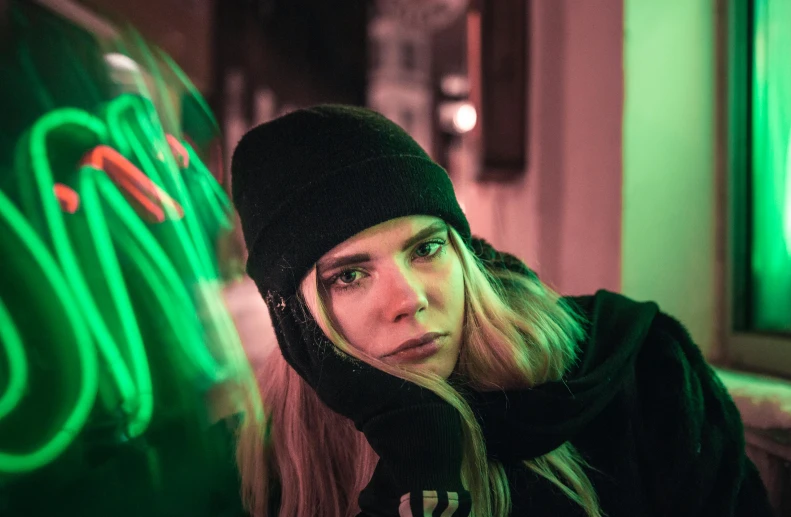 a woman standing in front of a neon sign, a character portrait, inspired by Elsa Bleda, trending on pexels, realism, black beanie, green glowing eyes, emma watson with anxious, blonde swedish woman