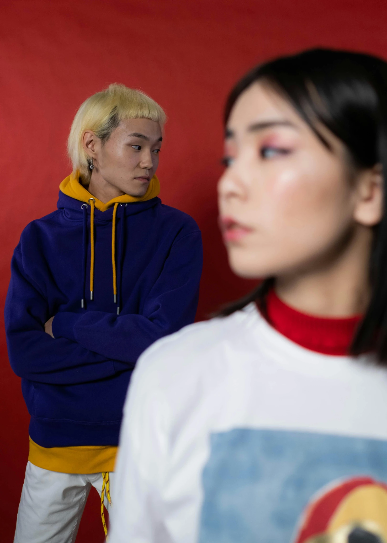 two people standing next to each other in front of a red wall, inspired by Tadanori Yokoo, trending on pexels, realism, young man in a purple hoodie, yellow and blue, portrait androgynous girl, ethnicity : japanese