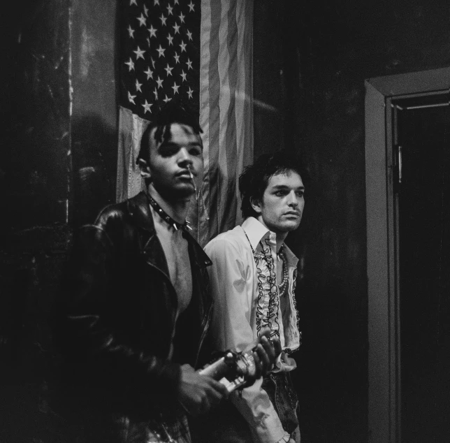 two men standing next to each other in front of an american flag, a black and white photo, by Maurycy Gottlieb, unsplash, renaissance, a 1980s goth nightclub in soho, young prince, playboi carti portrait, badlands