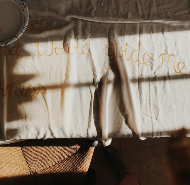 a pillow sitting on top of a bed next to a window, a silk screen, inspired by Tracey Emin, pexels contest winner, sitting on a mocha-colored table, gold and white cloak, sun flares, white apron