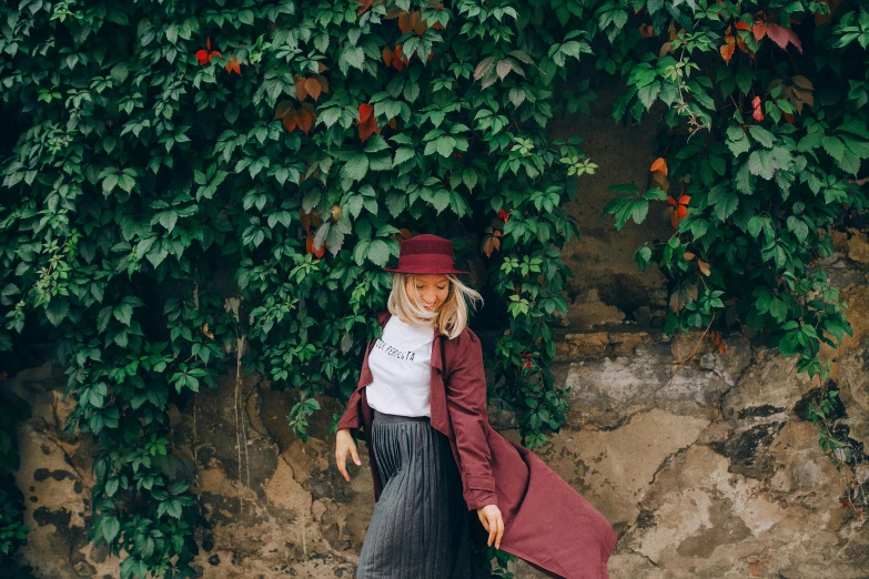 a woman standing in front of a stone wall, a photo, by Emma Andijewska, pexels contest winner, graffiti, maroon hat, long skirt, foliage clothing, wearing a trenchcoat
