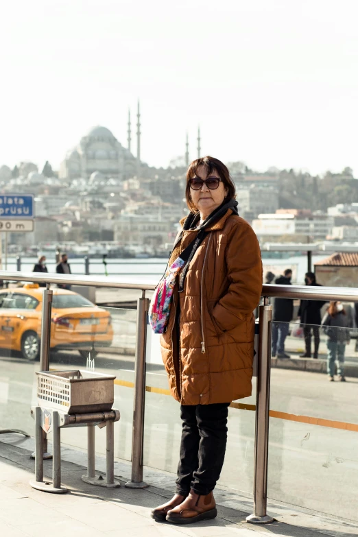 a woman standing on a sidewalk next to a railing, a picture, hurufiyya, turkish and russian, city backdrop, 5 5 yo, brown
