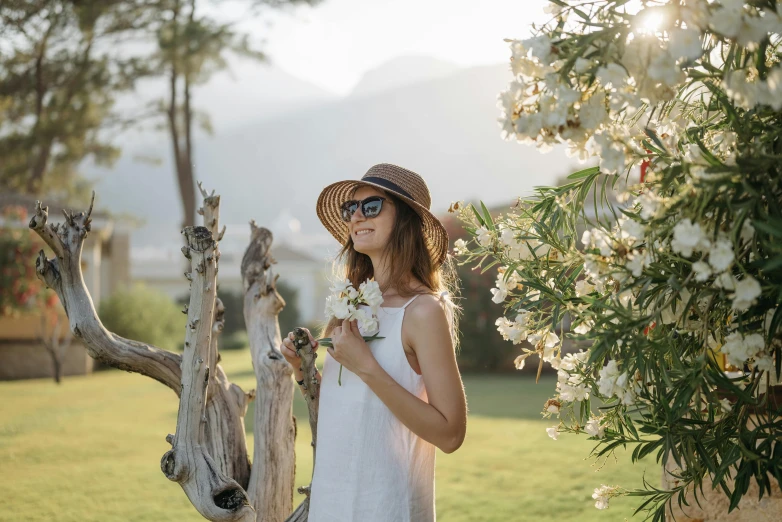 a woman standing next to a tree wearing a hat and sunglasses, pexels contest winner, white sundress, avatar image, picking flowers, mountains in the background