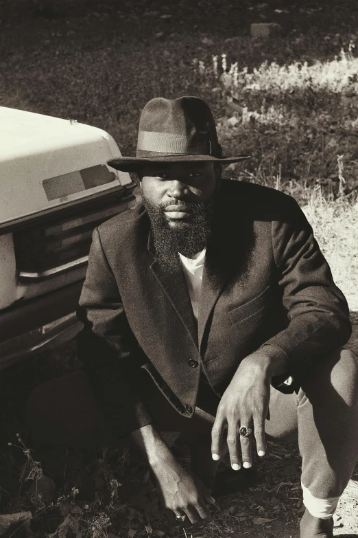 a man sitting on the ground next to a car, an album cover, by David Eugene Henry, mc ride, wearing a fedora, 1990 photograph, thick black beard