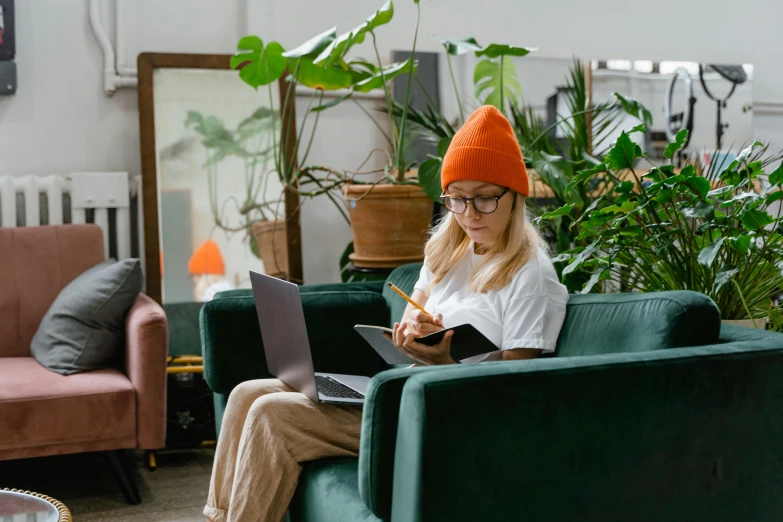 a woman sitting on a couch with a laptop, trending on pexels, green hat, terrarium lounge area, orange balaclava, student